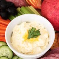 Cream Cheese Veggie Dip in a white bowl with garnish surrounded by cucumbers, carrots, celery, salami, apples and grapes.