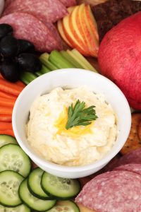 Cream Cheese Veggie Dip in a white bowl with garnish surrounded by cucumbers, carrots, celery, salami, apples and grapes.
