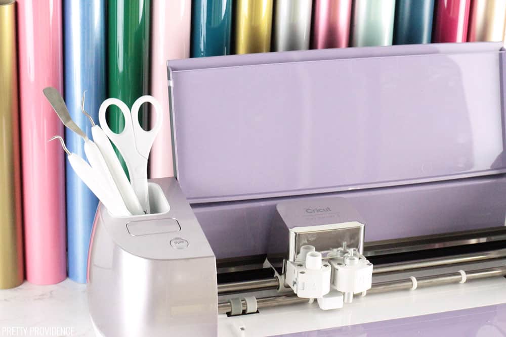 Cricut Explore Air 2 Wisteria (lilac) with cricut tools in and rolls of vinyl behind it.