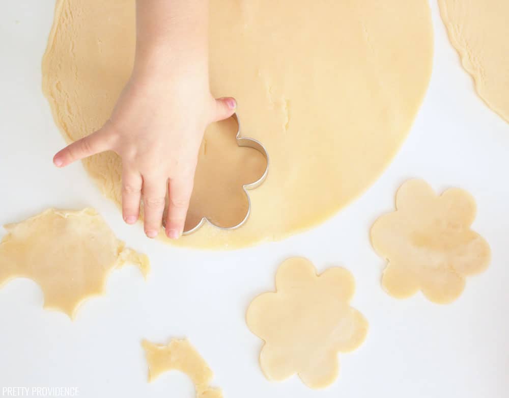 Pie crust rolled out on a white surface, a small hand cutting out pie crust with a cookie cutter.