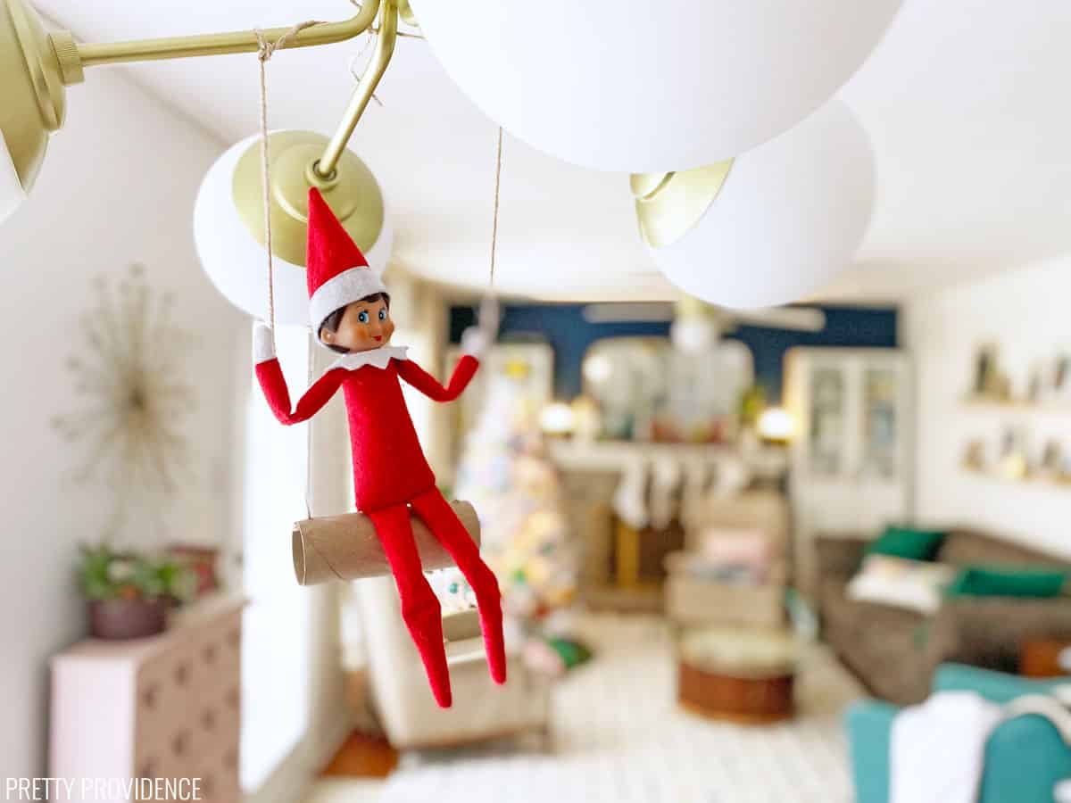 Elf on the Shelf sitting on a swing made from a toilet paper roll and twine, hanging up on a light fixture.