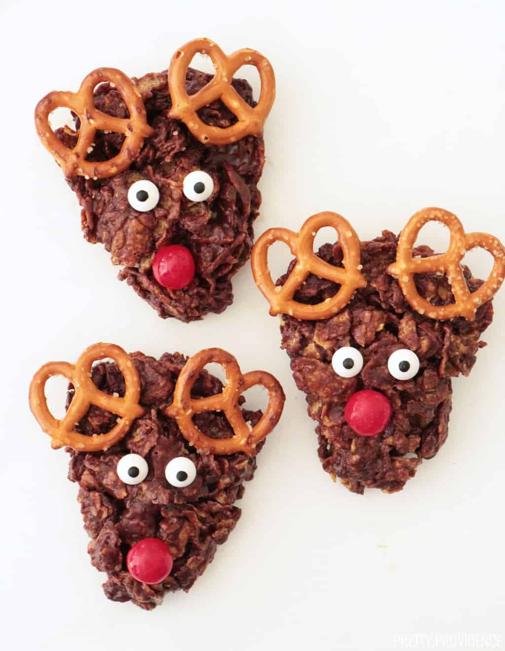 Three reindeer cookies made with cornflakes, marshmallows, pretzels, chocolate and candies.