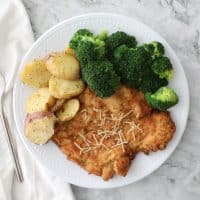 chicken schnitzel, broccoli and potatoes on a white plate on a granite counter with a white napkin on the left