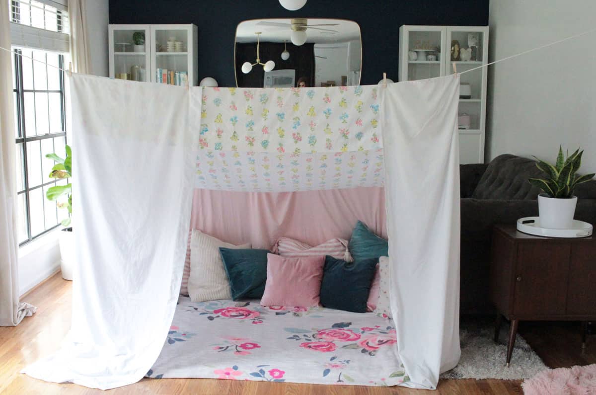 Canopy blanket fort living room tent made from white bed sheets, pillows, blankets and held up by clothes line, clothes pins and chairs.