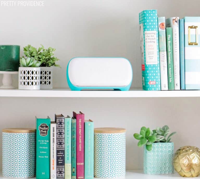 Cricut Joy machine on a bookshelf between books and plants.