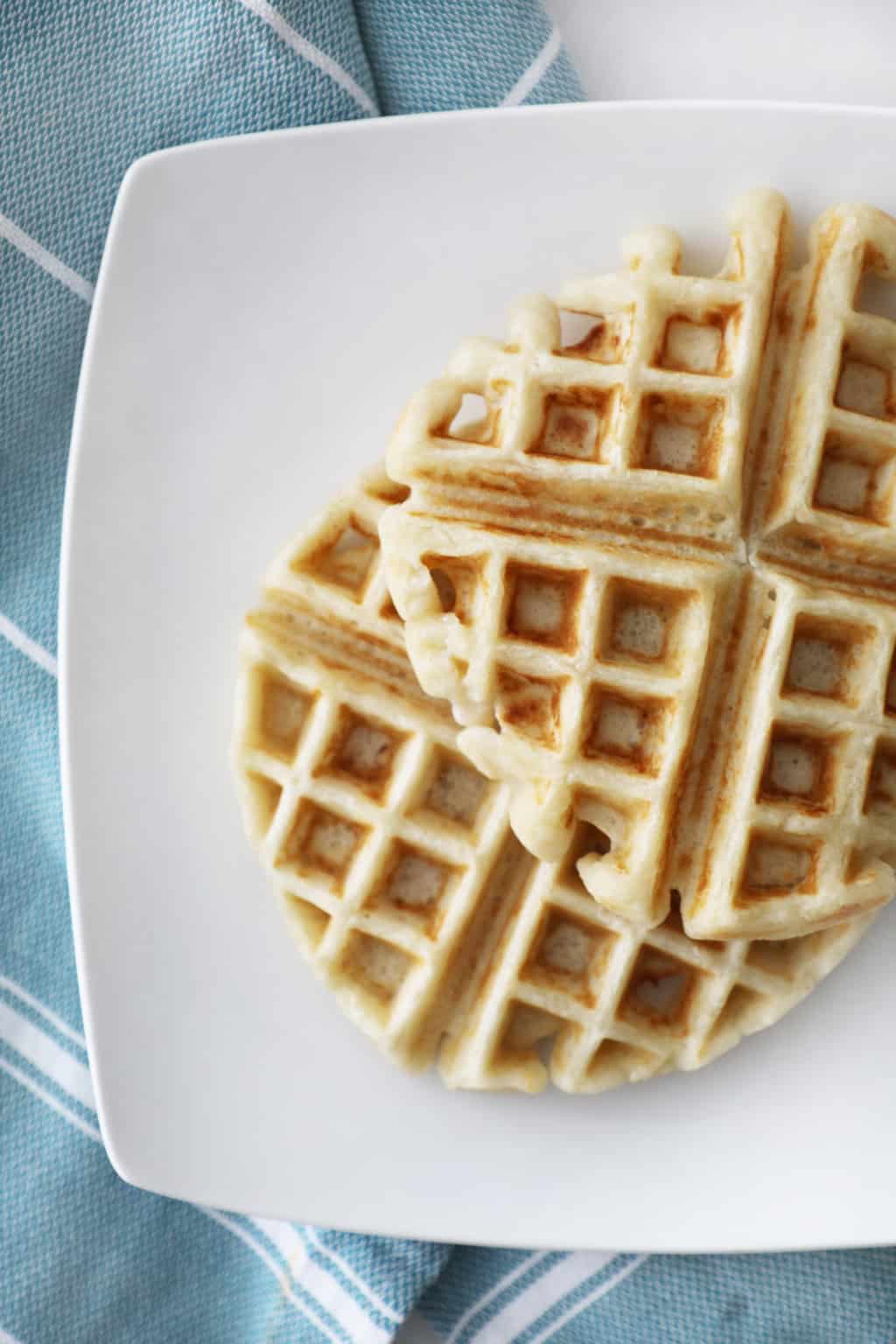 two plain yeast waffles on a white square plate next to a blue tea towel
