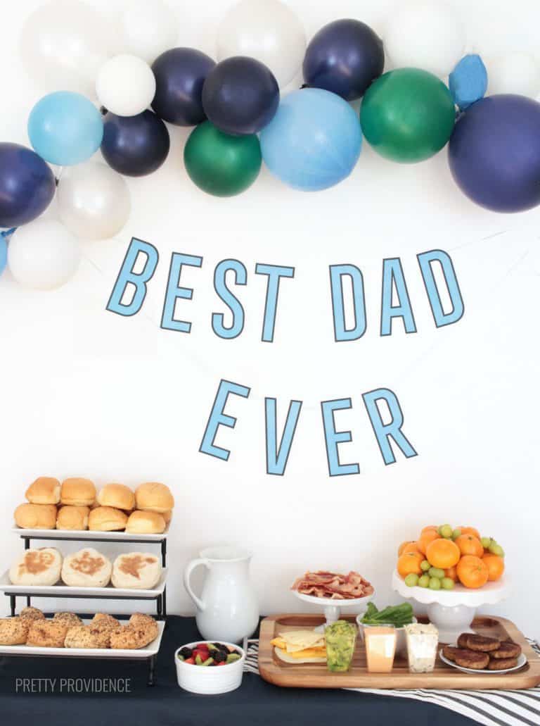 Father's Day breakfast table with breakfast sandwich ingredients, balloons and 'best dad ever' banner.