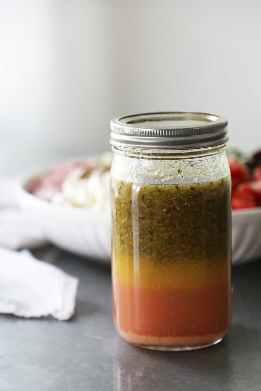 homemade italian dressing in a mason jar on a cement table with a white napkin and bowl of pasta in the background