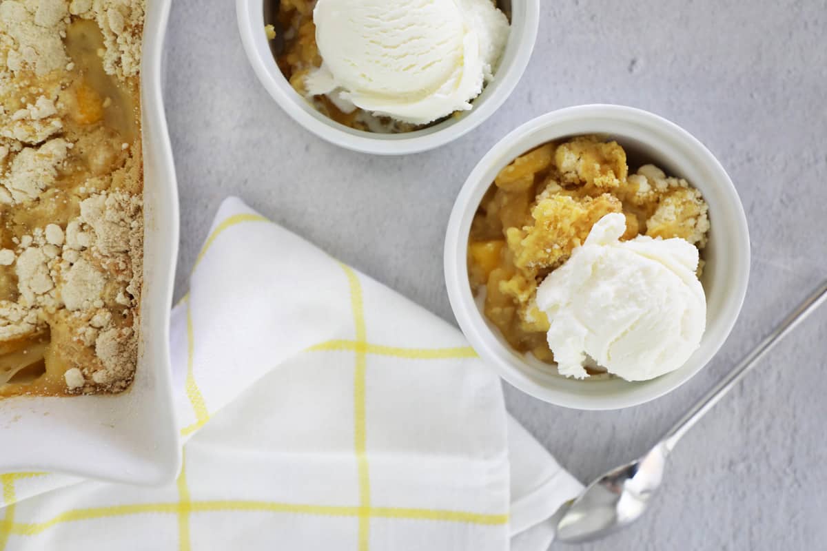 two dishes of peach cobbler with ice cream next to the full pan and a white towel