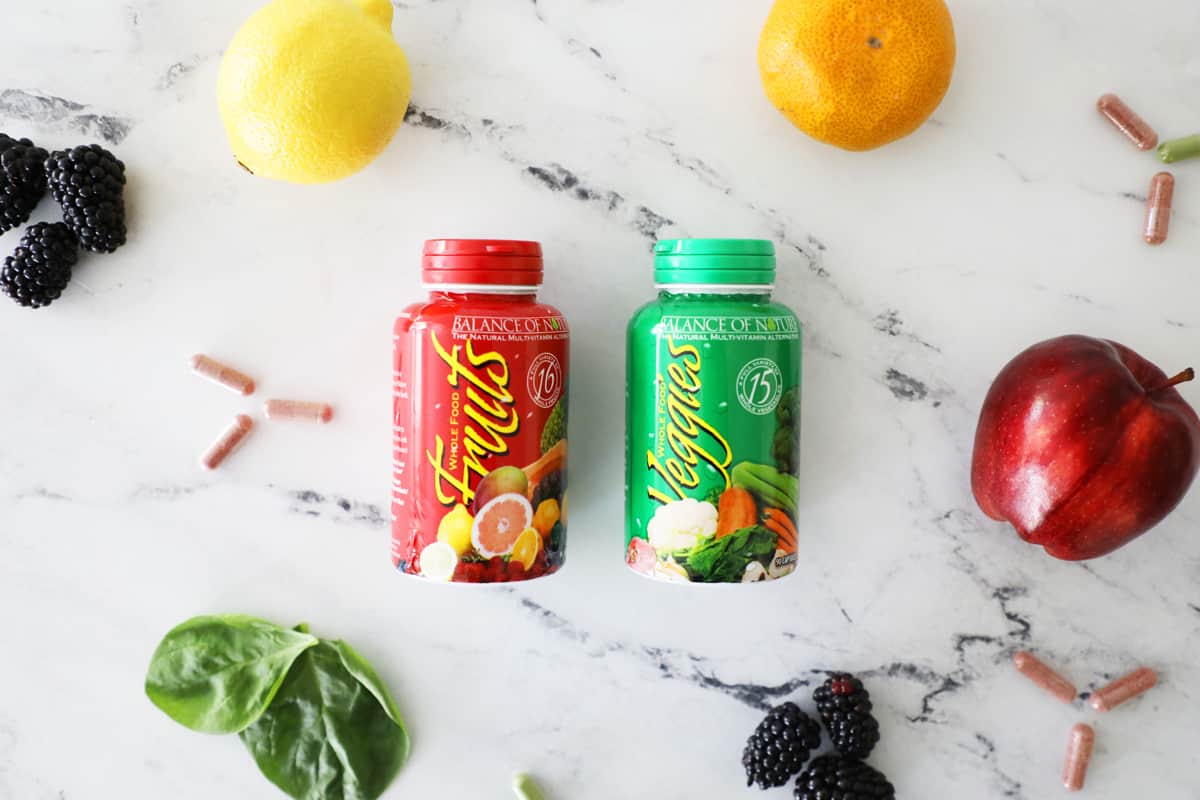 fruit and veggie capsules on a white marble countertop surrounded by fruit and supplements