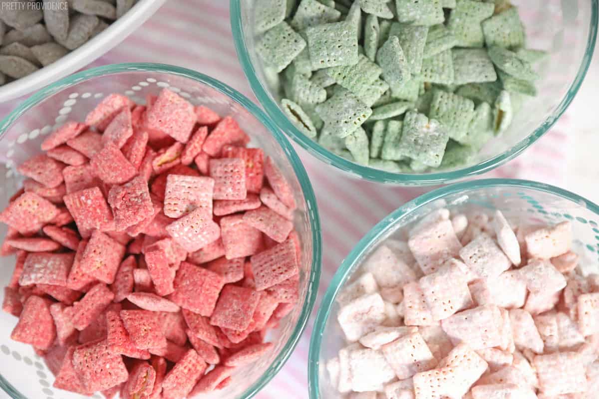 Chex cereal covered in red, pink and green candy coating and powdered sugar separated into bowls by color, muddy buddies AKA puppy chow