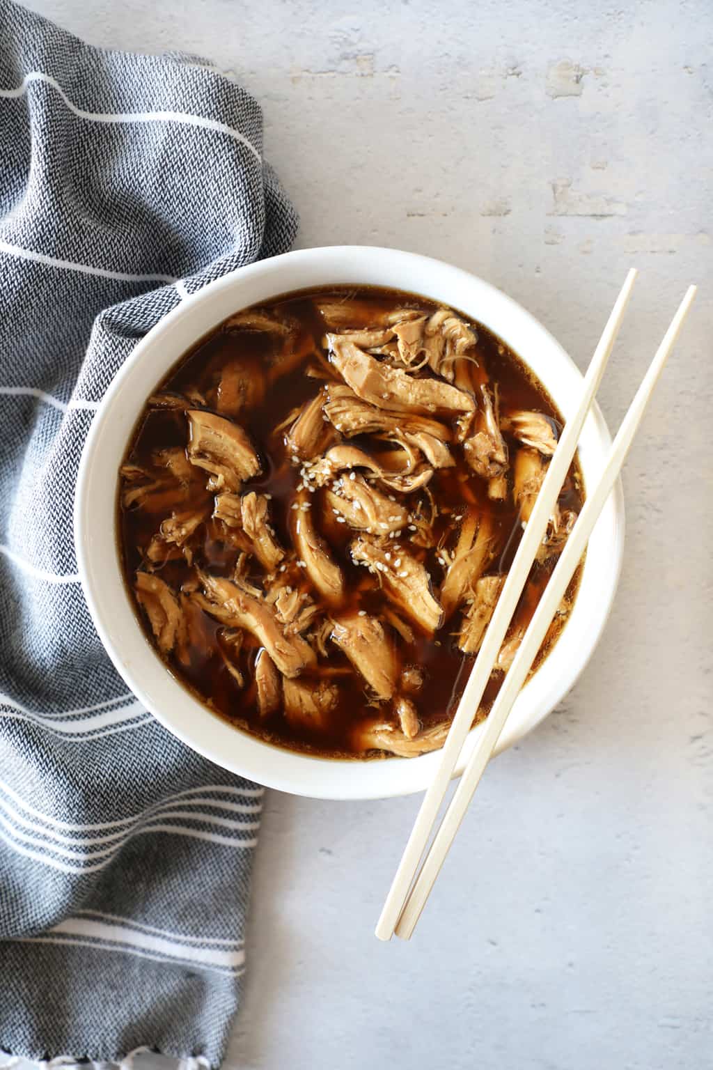 teriyaki chicken in a white bowl next to a grey tea towel and chopsticks