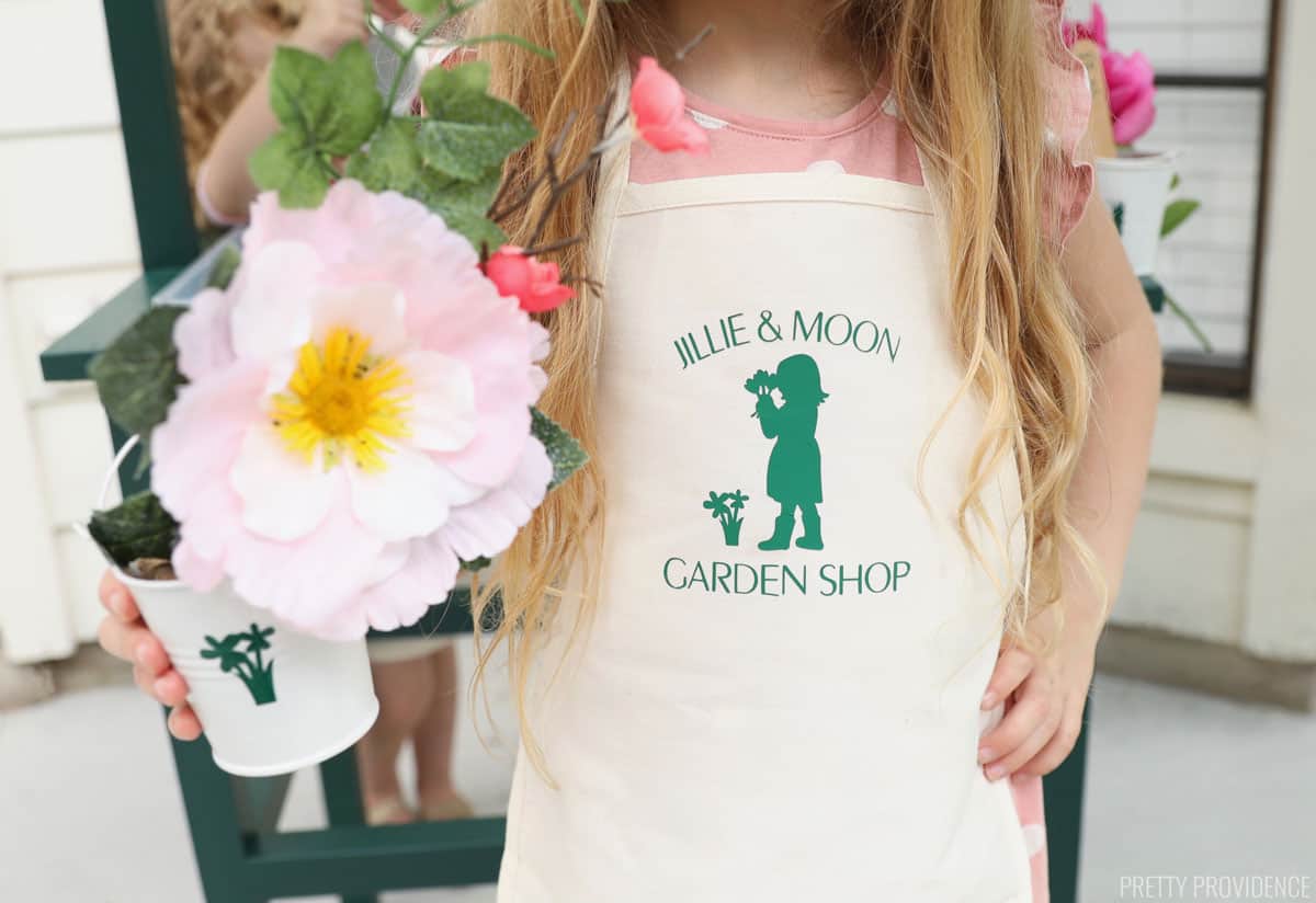 little girl holding flowers and wearing an apron with flowers and garden shop logo on it