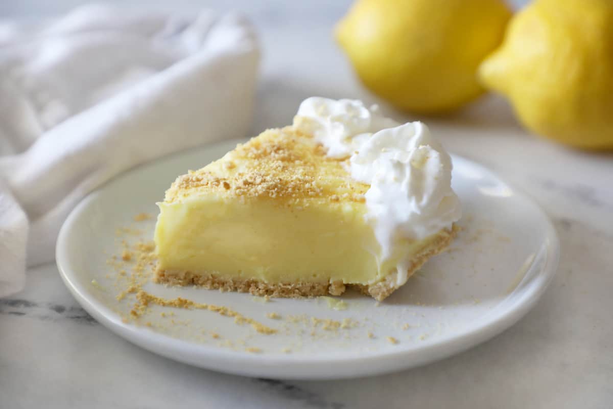 a slice of lemon pudding pie on a white plate next to a white cloth napkin