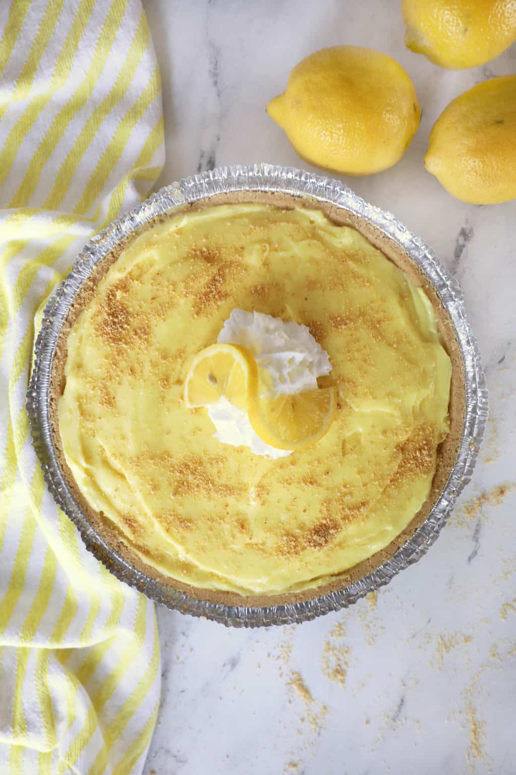 a full lemon pudding pie next to a towel, some lemons and crumbs