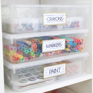Three clear bins with markers, paint and crayons in them, with white labels