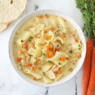 Chicken noodle soup with diced carrots, diced celery, shredded chicken and egg noodles in a white bowl, with carrots and bread next to it.