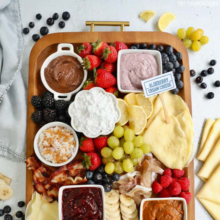 Breakfast charcuterie board with crepe toppings, fruit, spreads, bacon and powdered sugar
