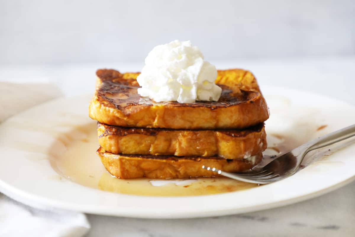 a stack of pumpkin French toast with whipped cream and syrup and a fork next to it