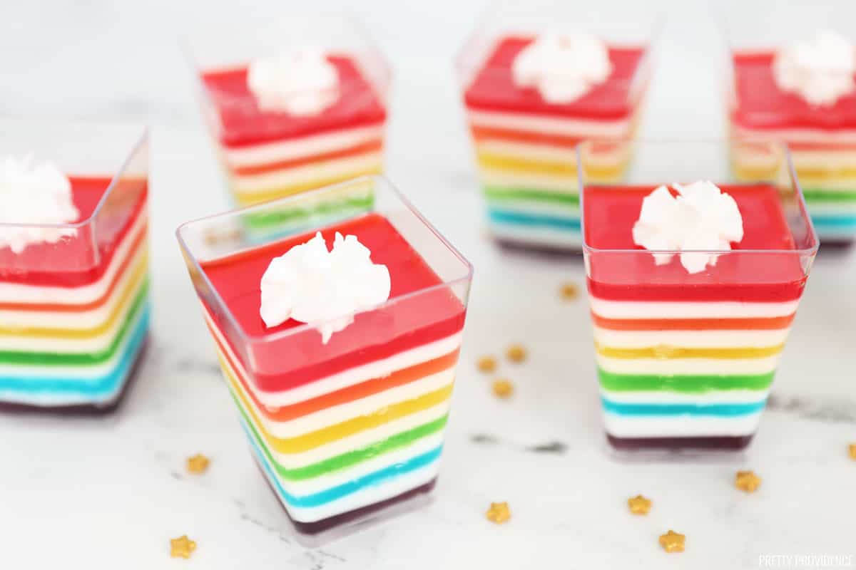 Rainbow jello layered in clear, square cups with a dollop of whipped cream on tp