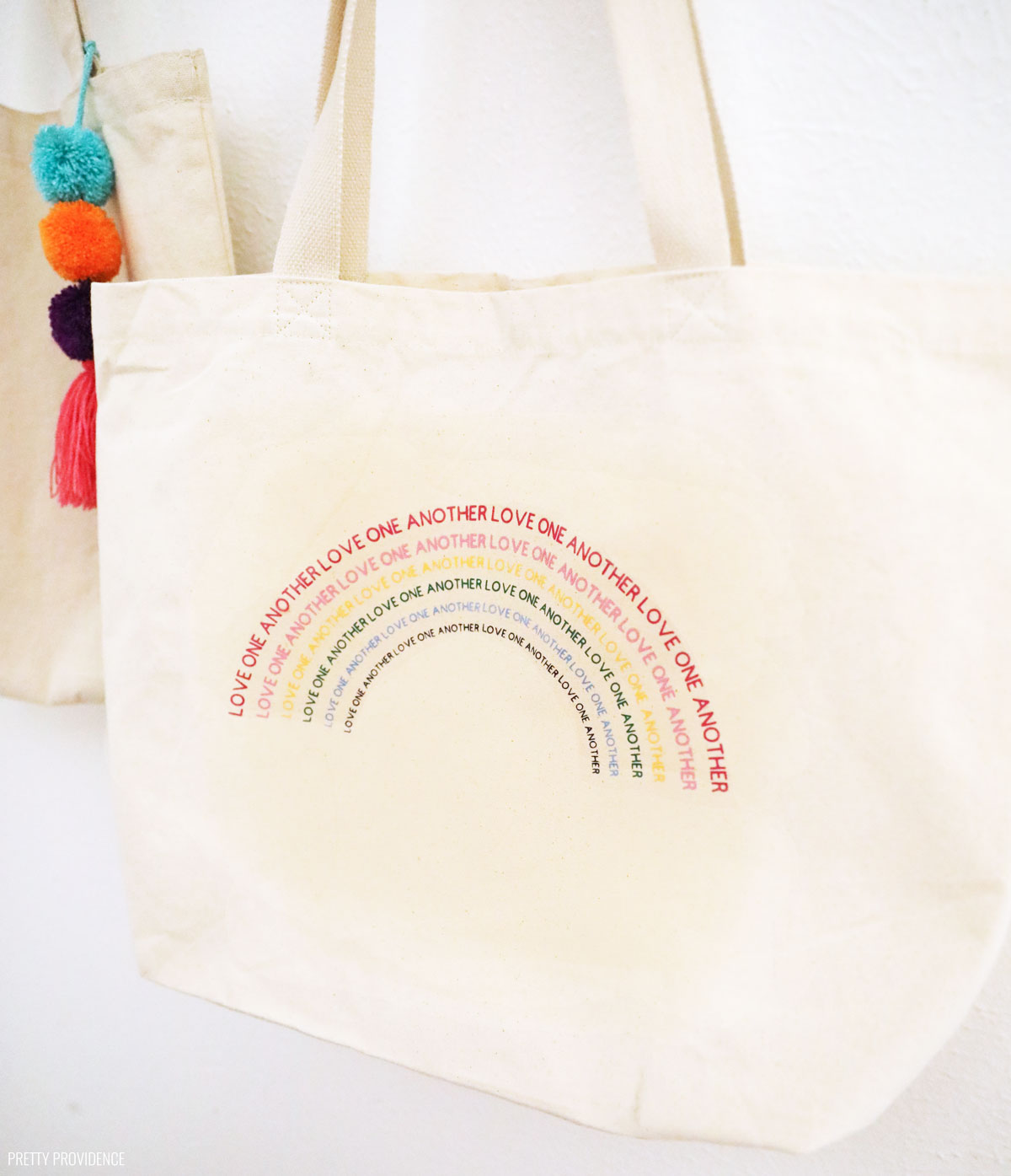 love one another words forming a rainbow on a tote bag hanging up against a white wall