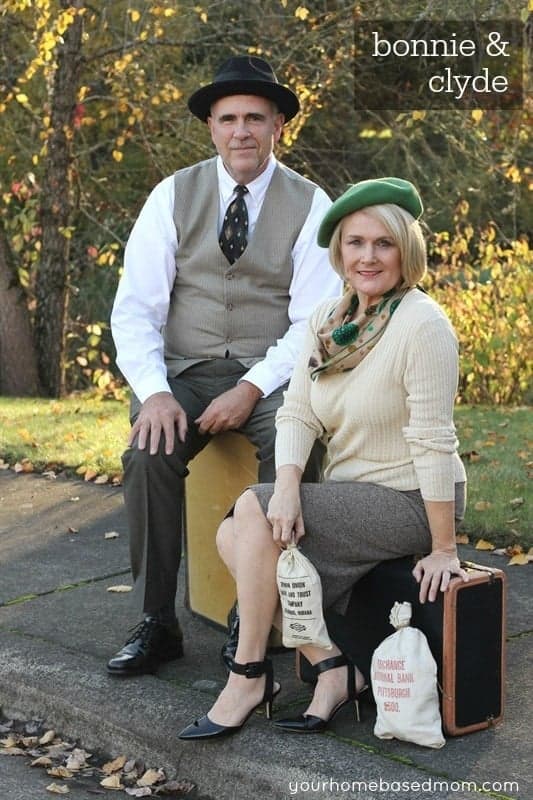 A woman and man dressed as Bonnie & Clyde costume as a couples Halloween costume sitting on suitcases with bags of money by their feet.