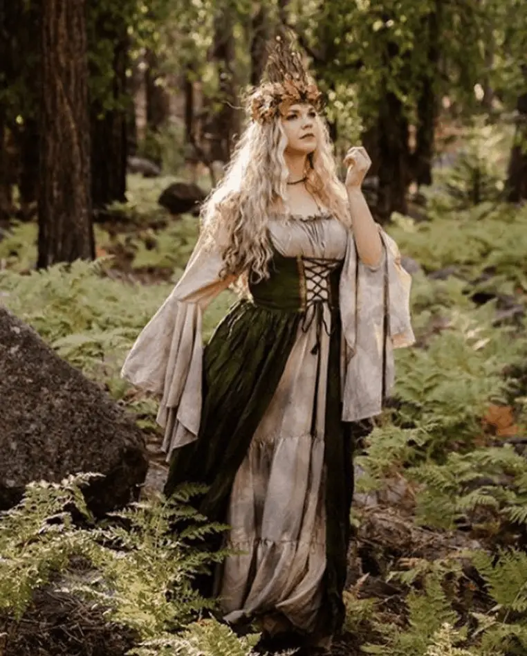 A woman in the forest wearing a forest fairy costume. She is wearing a medieval-style dress and a crown made from leaves, feathers and flora.