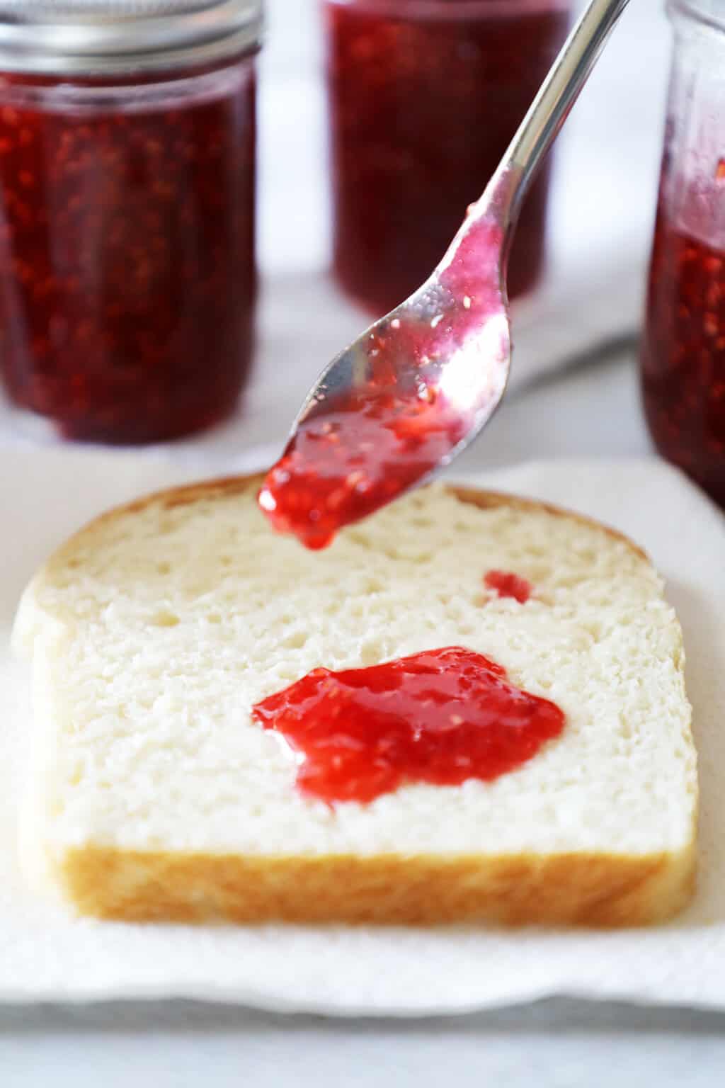 a silver spoon with raspberry jam on it hovering over and dripping onto a piece of white bread