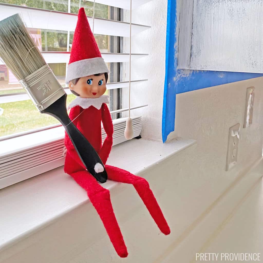 Elf on the shelf with a paintbrush on a windowsill next to a wall with painter's tape on it..