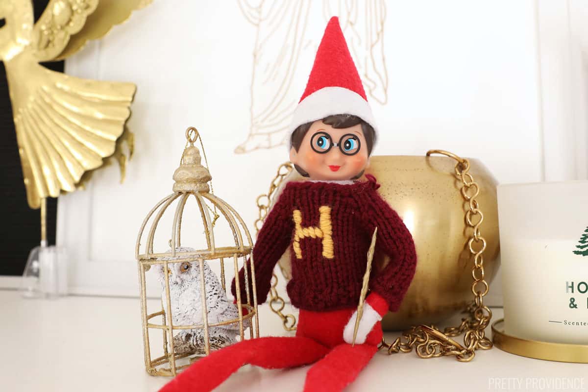 Elf on the Shelf dressed as Harry Potter -wearing a crimson sweater with a gold H on it, holding a wand and sitting next to a snowy owl. 