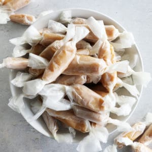 A white bowl overflowing with homemade caramels wrapped in parchment paper.