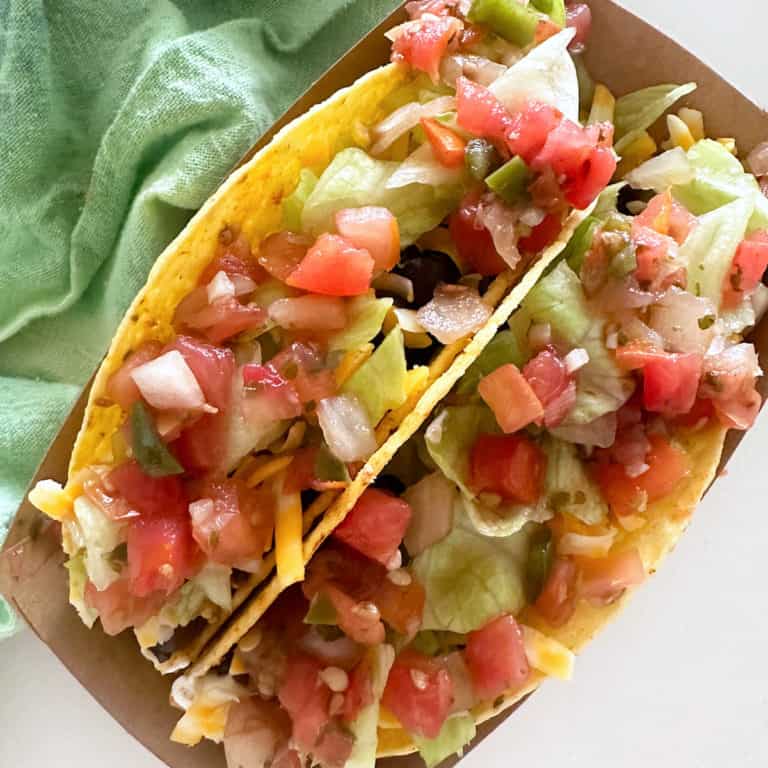 Two hardshell chicken tacos in a brown taco holder next to a green towel.