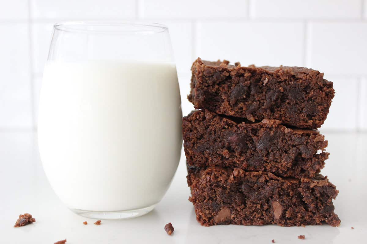 A glass of milk on a white countertop next to three homemade brownies in a stack.