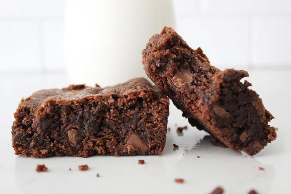 Two homemade brownies, one flat on the counter, the other tipped on top of the first with a glass of milk behind them.