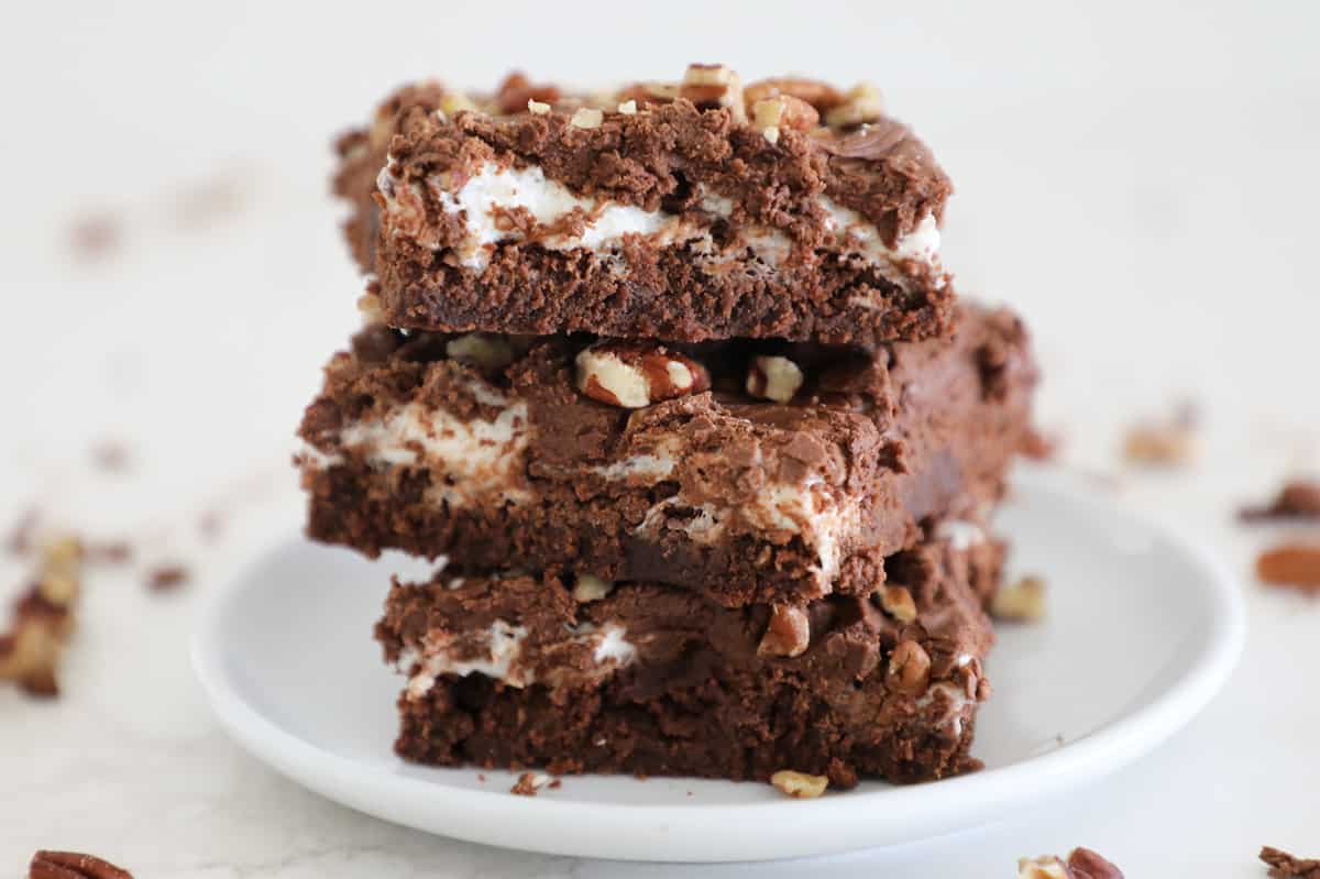 Three rocky road brownies stacked on a small white plate with pecans around it.