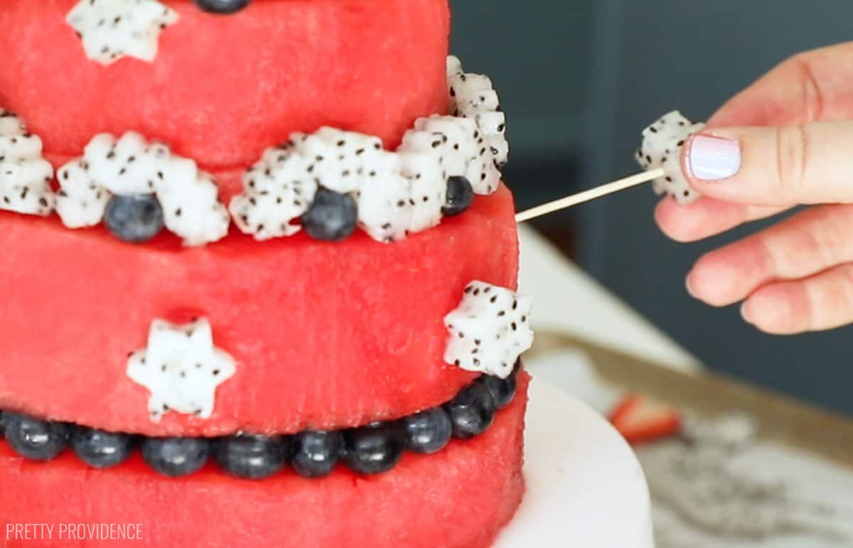 Fresh watermelon cake decorated with dragon fruit and berries, a toothpick being pushed into the take to hold in place.