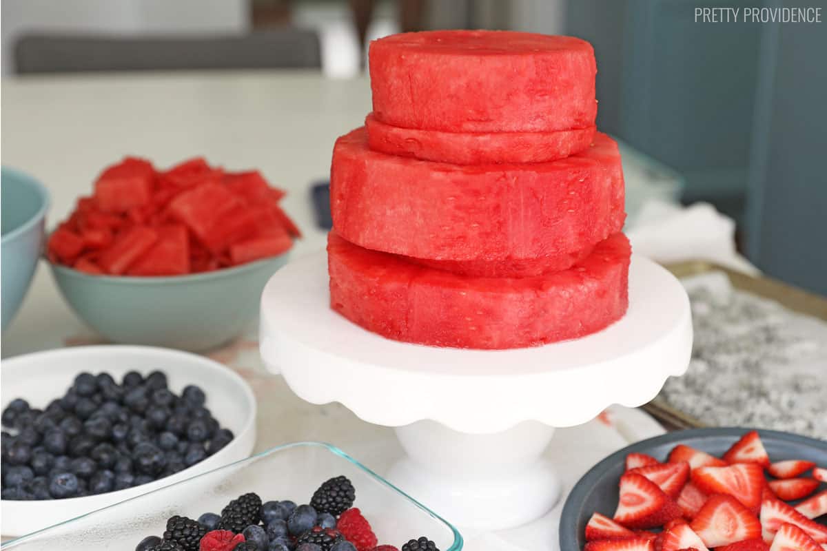 Fresh watermelon cut into a two-tier cake shape on a white cake stand, surrounded by fresh berries..