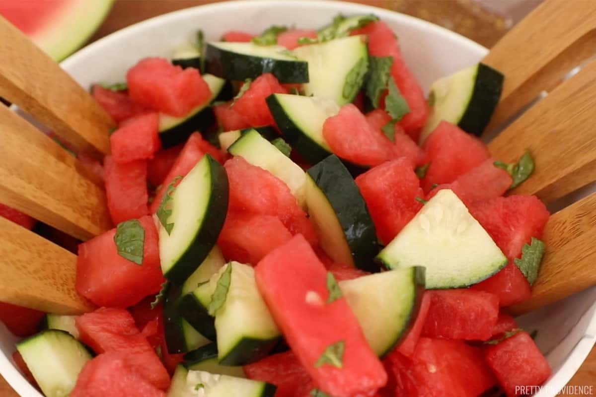 watermelon cubes, cucumber slices and mint leaves being mixed together with salad servers in a white bowl. 
