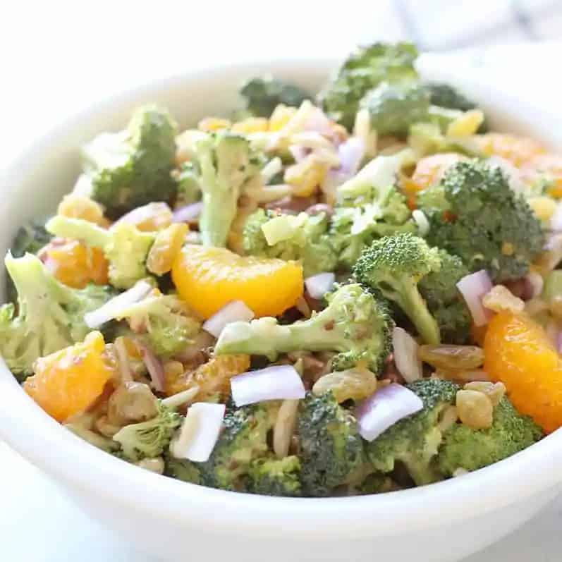 Broccoli salad with mandarin oranges, red onion, slivered almonds and golden raisins in a white bowl.