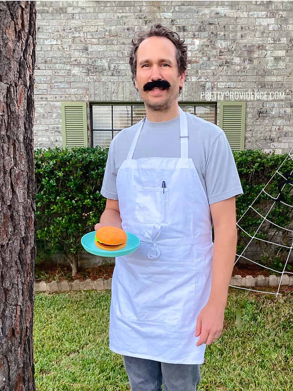 Man dressed as Bob Belcher from Bob's Burgers Costume wearing a gray t-shirt, white apron, bushy black mustache and holding a toy hamburger on a plate. 