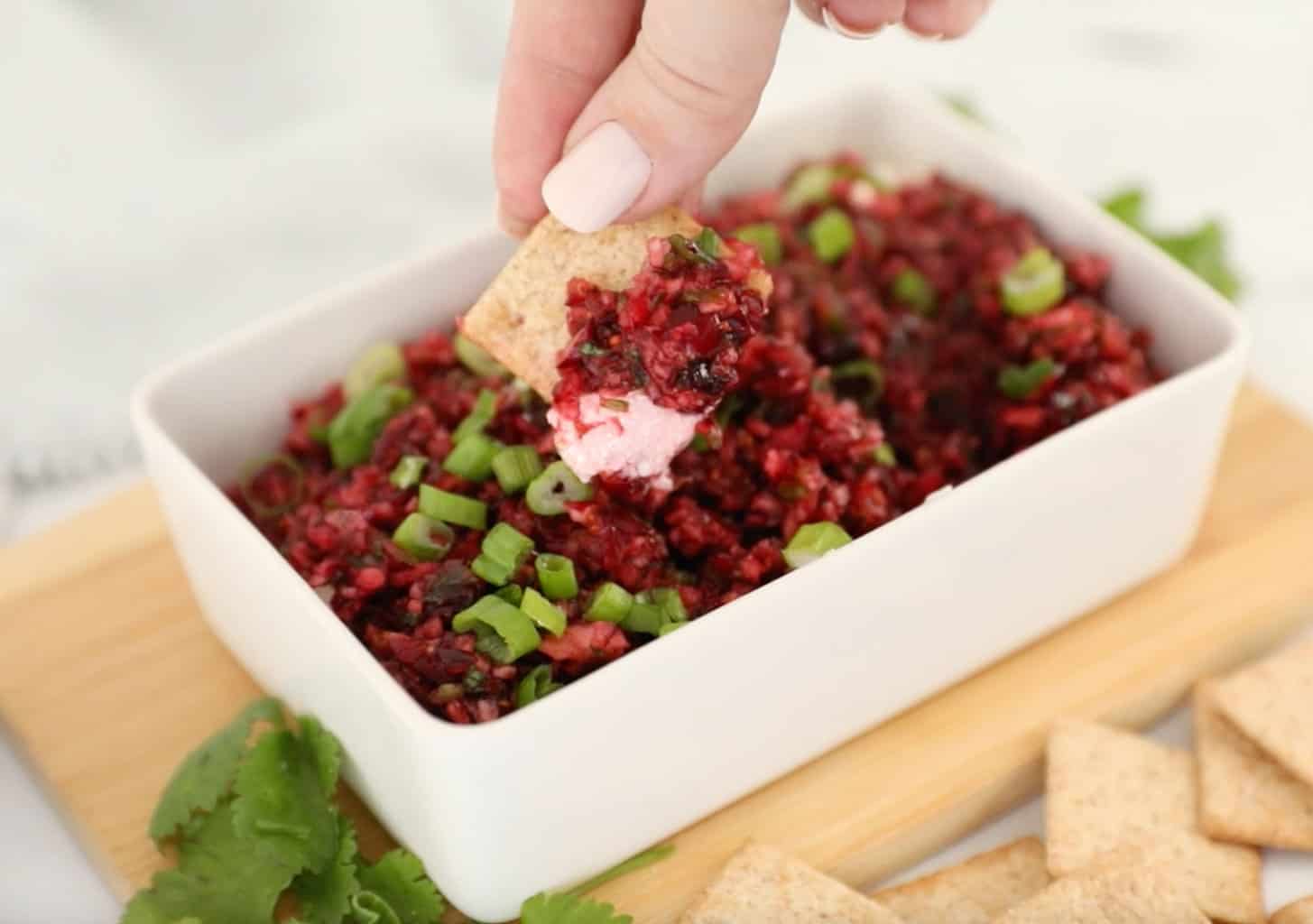 Cranberry Jalapeño Dip with cream cheese in a white dish with a hand lifting a cracker with a scoop of dip on it out of the dish.