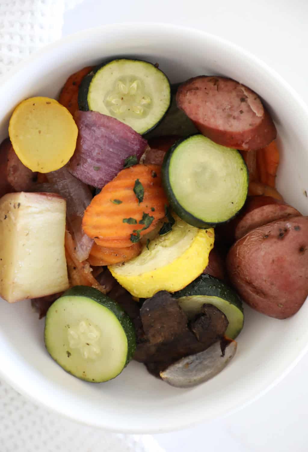 Roasted kielbasa and veggies in a white bowl on a white countertop.