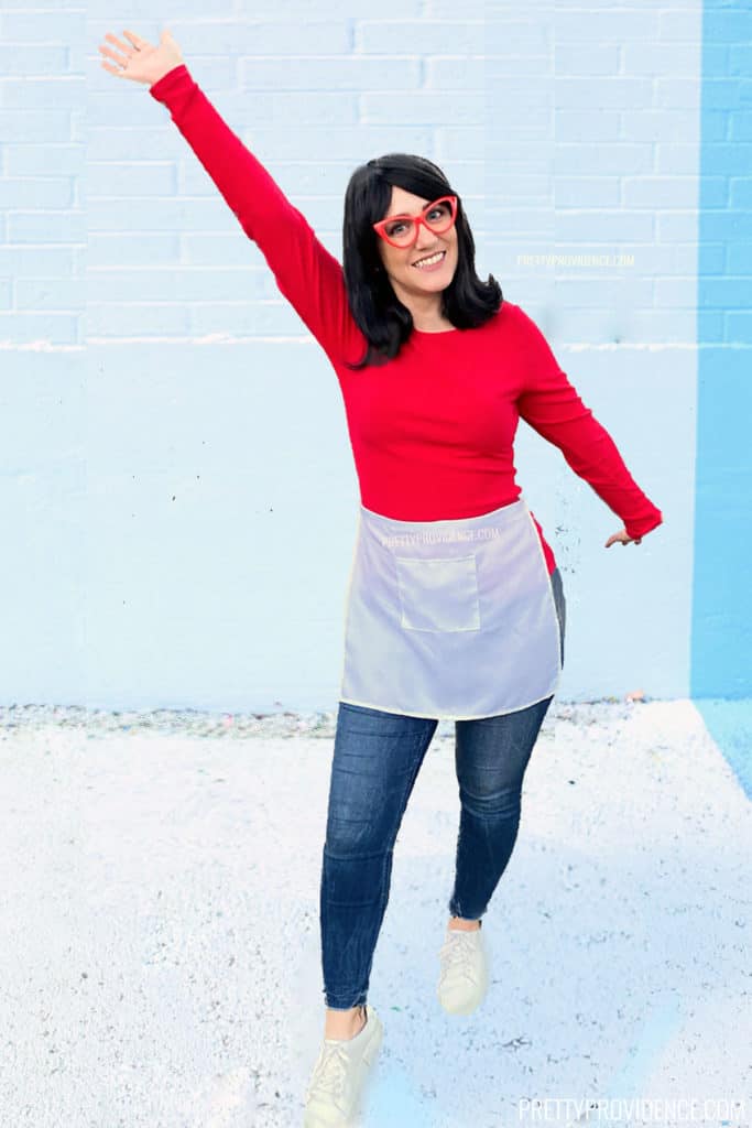 Linda Belcher from Bob's Burgers costume on a woman with a red shirt, white half-apron, red glasses and black wig.