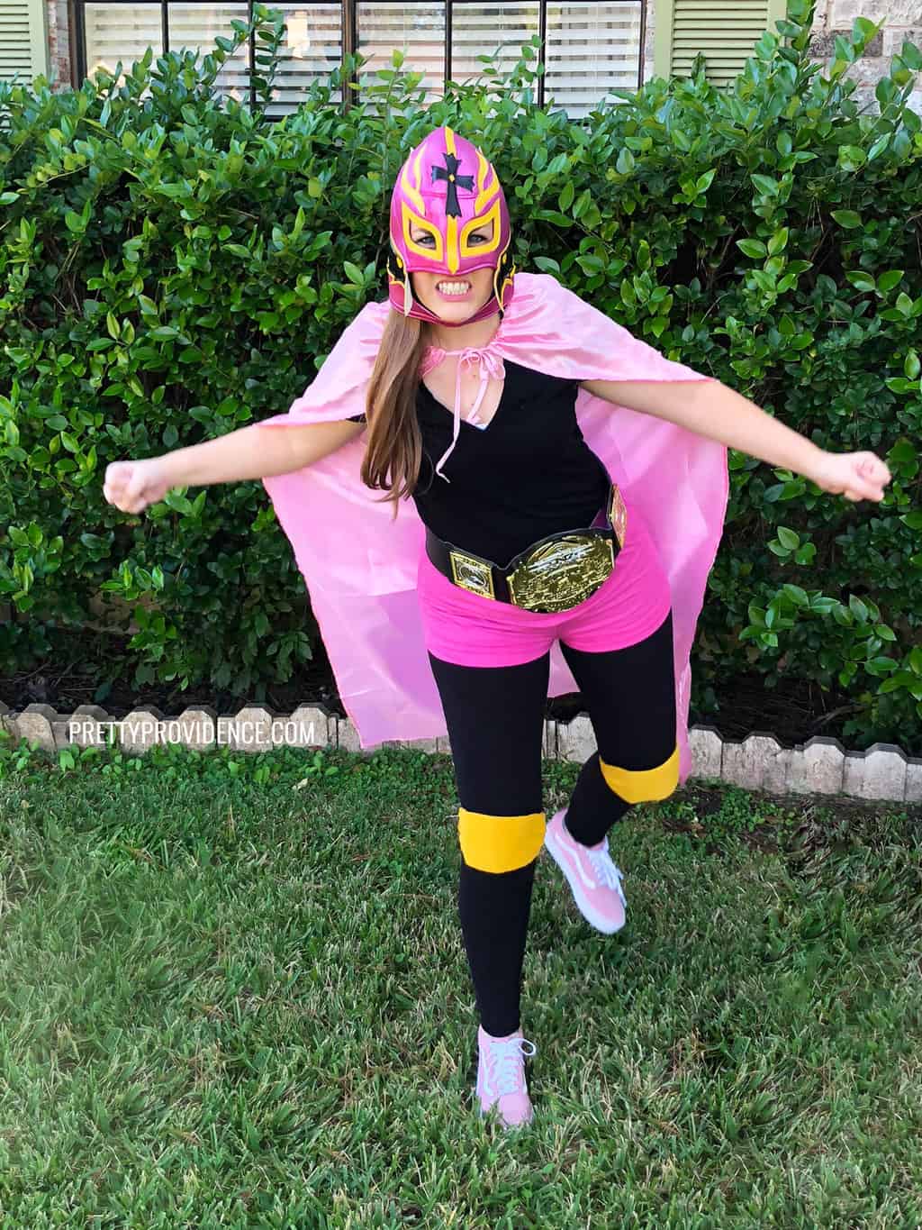 Woman wearing a lucha libre costume with a pink and yellow luchador mask, a pink cape, pink shorts, black wrestling belt and yellow knee pads.