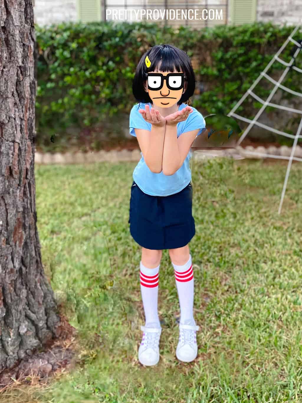 A kid dressed as Tina fron Bob's Burgers wearing a blue t-shirt, navy blue skirt, tube socks, a bob haircut and a yellow hair clip.
