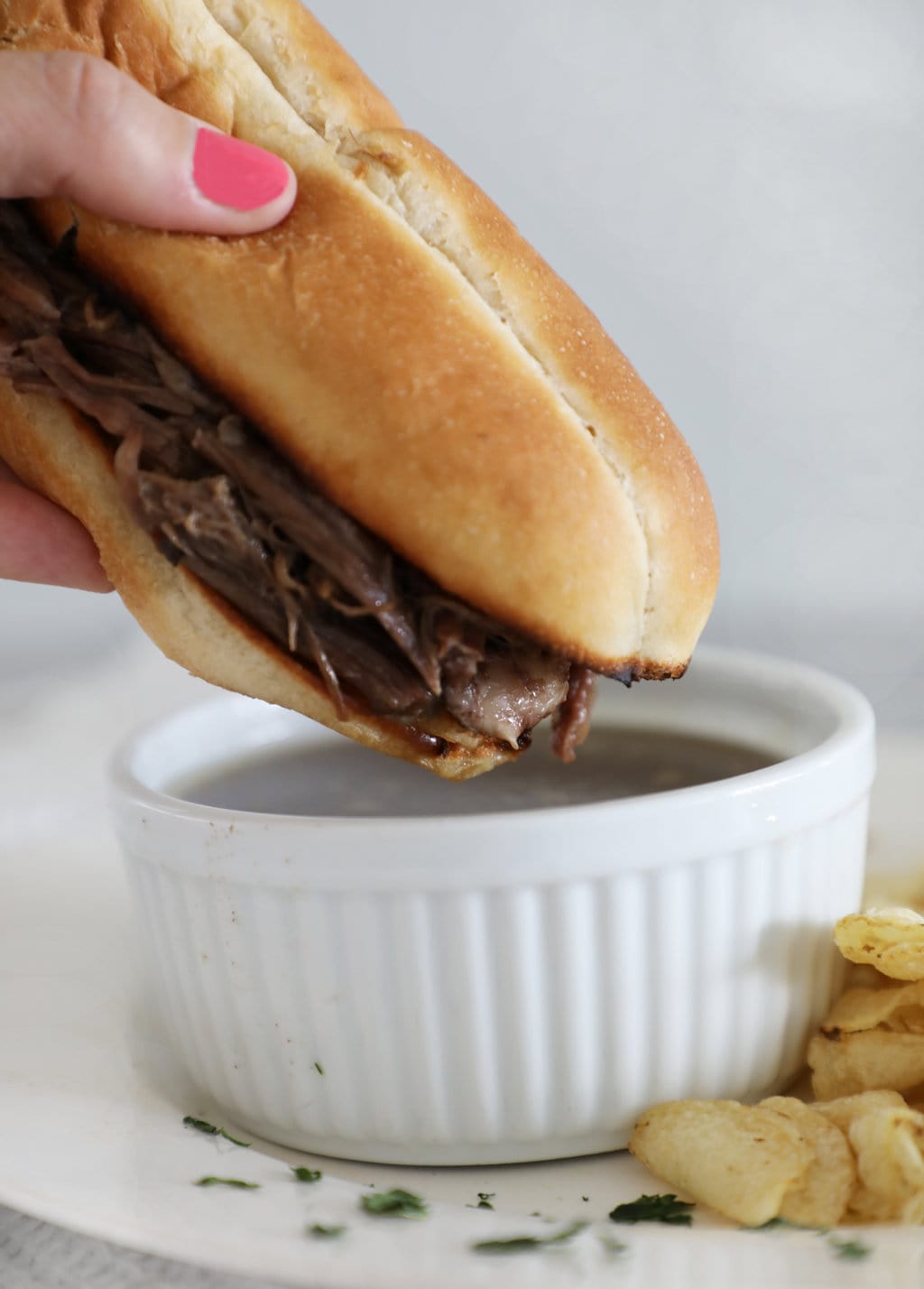 A hand with a pink thumbnail dipping a French dip sandwich into a bowl of au jus. 