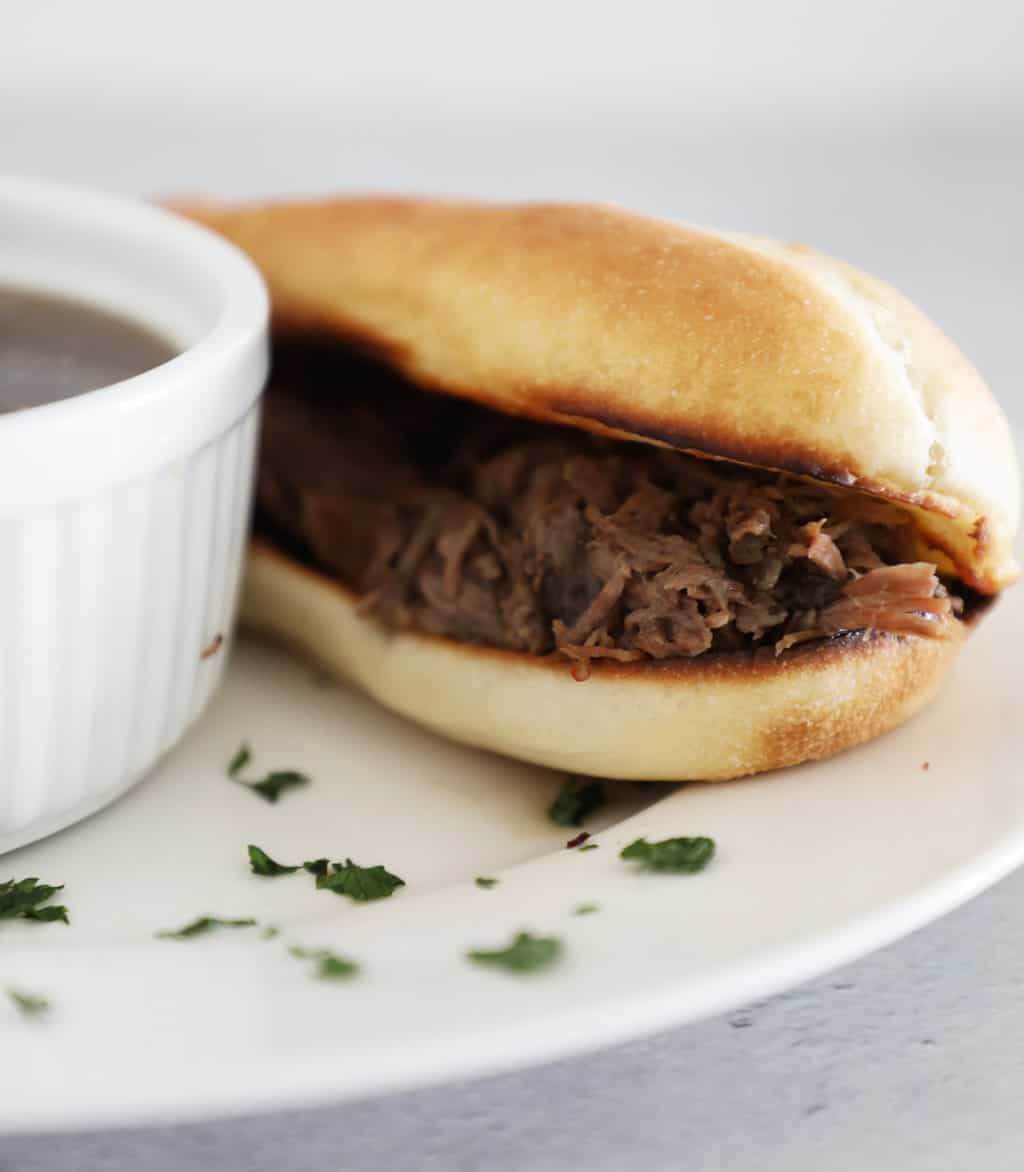 Side view of roast beef on a sandwich bun next to a bowl of au jus. 