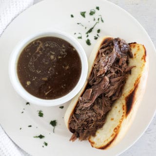 A white plate with a French dip sandwich next to a small bowl of au jus.