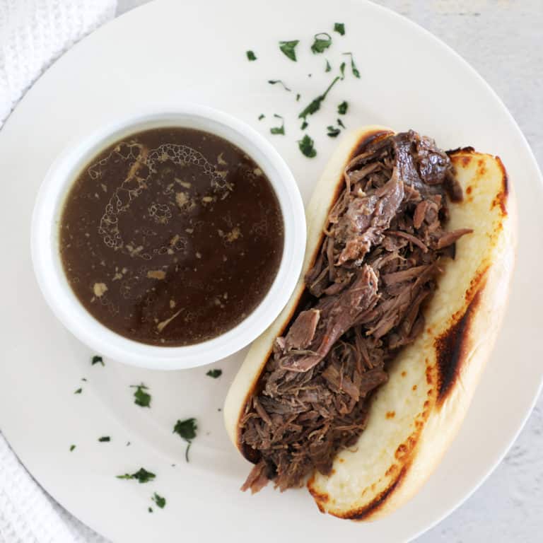 A white plate with a French dip sandwich next to a small bowl of au jus.