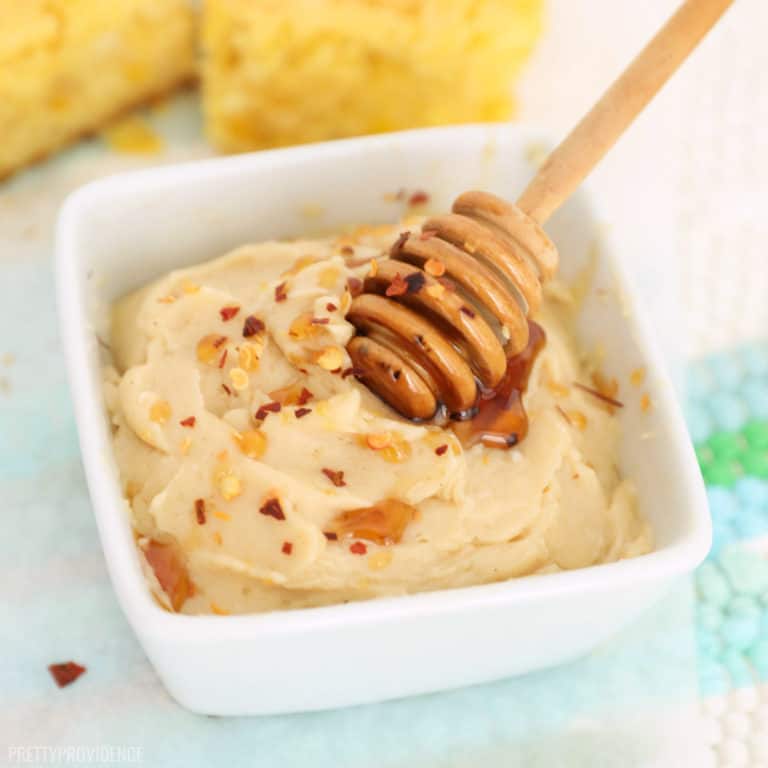 Honey butter in a small white dish with hot honey on a small honey wand and red pepper flakes sprinkled on top.